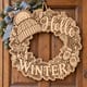 Unpainted wooden Hello Winter wreath with a knit hat and mittens hanging on a front door