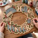 Hands painting the Hello Winter wreath with a knit hat and mittens at a craft table