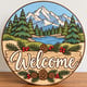 Painted round winter welcome sign with a mountain and pine tree scene standing on a wooden table
