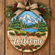 Painted round winter welcome sign with a mountain and pine tree scene hanging on a wooden front door