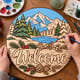 Hands painting the round winter welcome sign with a mountain and pine tree scene at a craft table