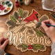 Hands painting the cardinal winter welcome door hanger at a craft table