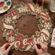 Hands painting the Tis the Season wreath with swirling ribbons and snowflakes at a craft table