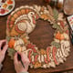 Hands painting the Thankful Thanksgiving wreath with a turkey and pumpkins at a craft table