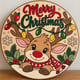 Painted round reindeer Merry Christmas sign with string lights standing on a wooden table