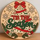 Painted round Tis the Season Christmas tree sign standing on a wooden table