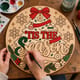 Hands painting the round Tis the Season Christmas tree sign at a craft table