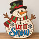 Painted Let It Snow snowman door hanger standing on a wooden table