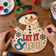 Hands painting the Let It Snow snowman door hanger at a craft table